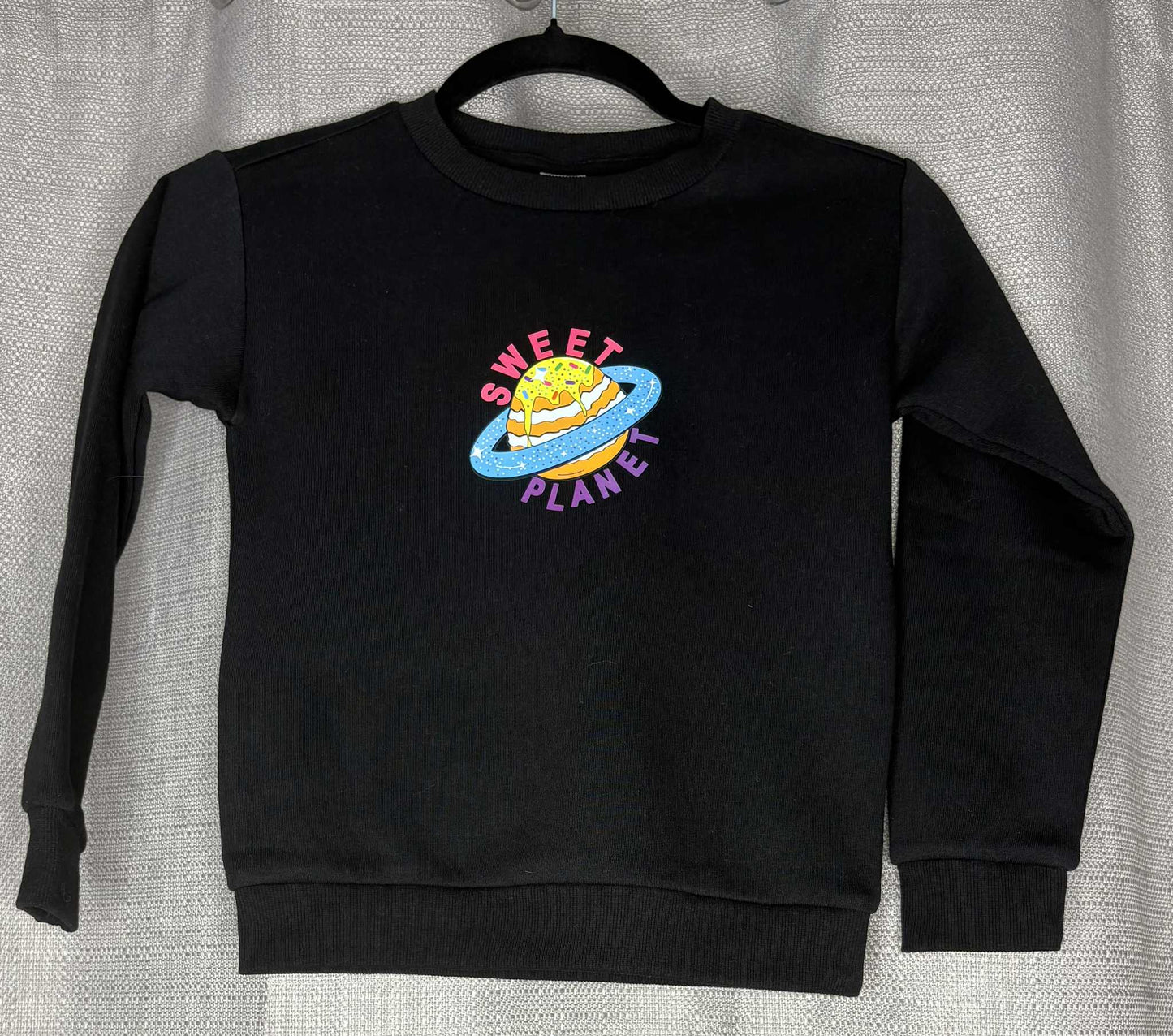 Black sweatshirt with 'Sweet Planet' graphic on a hanger against a neutral background