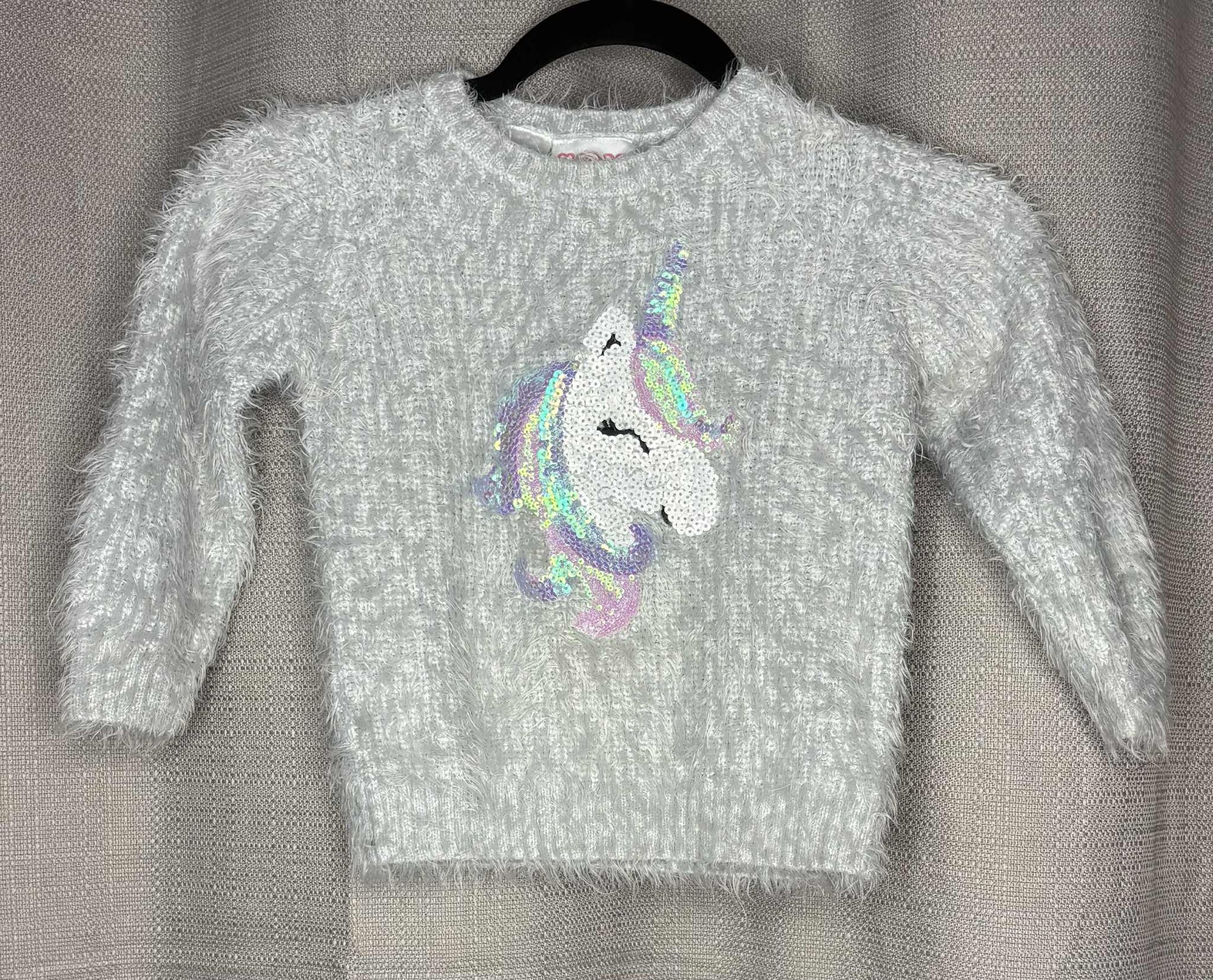 Gray sweater with a colorful unicorn design on a hanger against a gray background