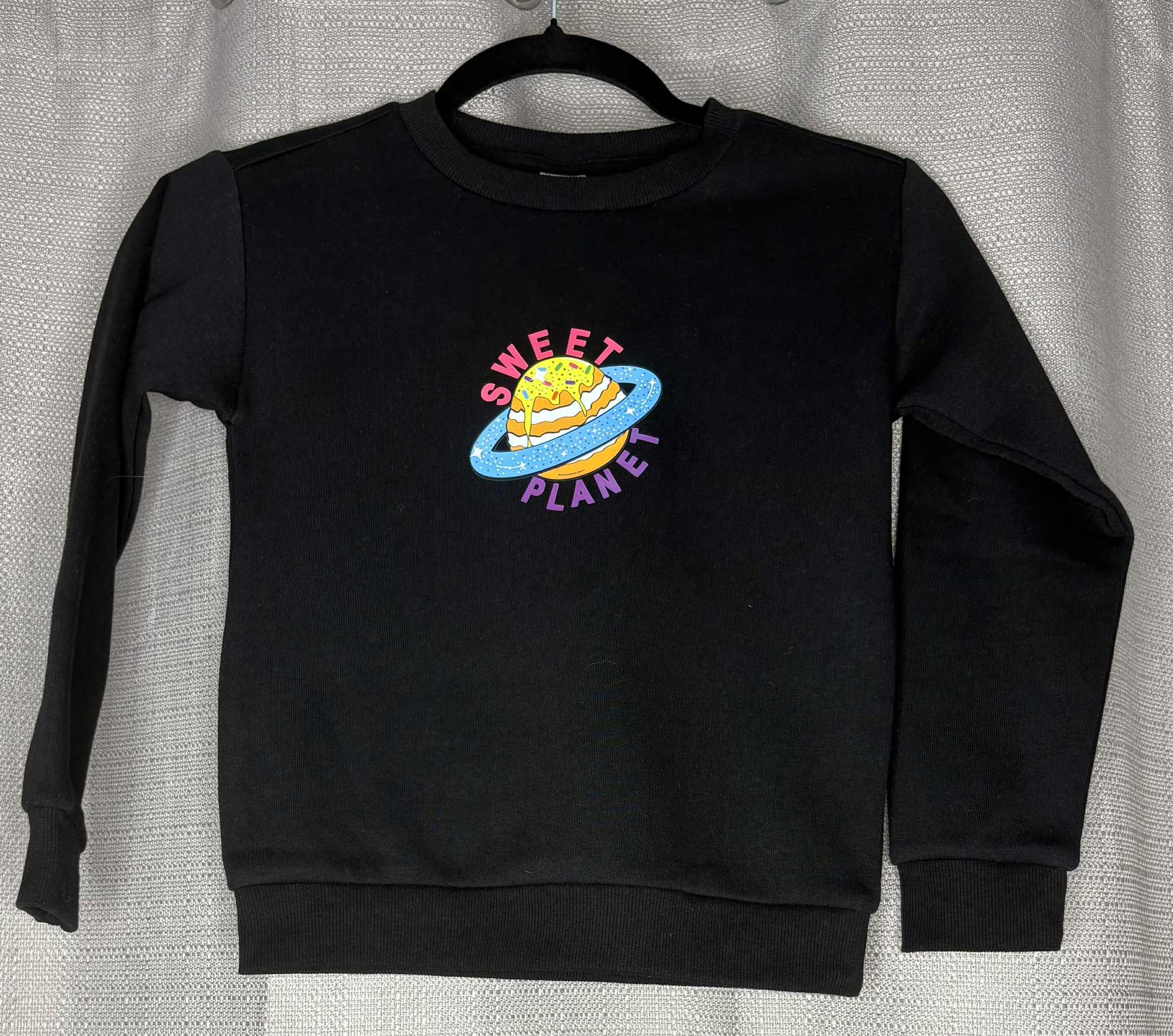 Black sweatshirt with 'Sweet Planet' graphic on a hanger against a neutral background